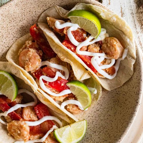 birds-eye view of shrimp tacos in corn tortillas on a white plate and topped with red bell peppers, lime slices and drizzled with plain greek yogurt