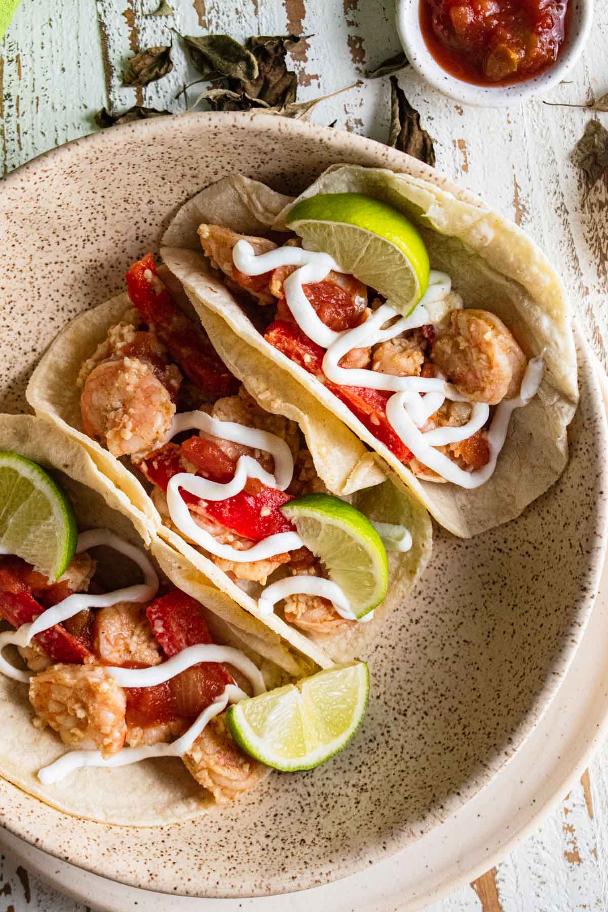 birds-eye view of shrimp tacos in corn tortillas on a white plate and topped with red bell peppers, lime slices and drizzled with plain greek yogurt