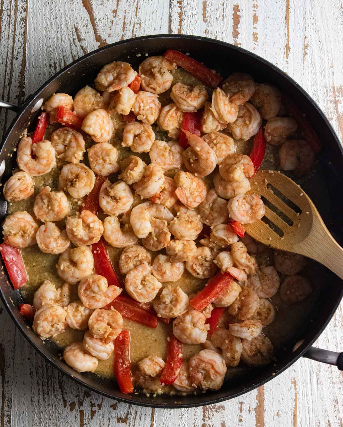 shrimp being cooked in a large non-stick skillet with red bell peppers being mixed with a wooden spoon