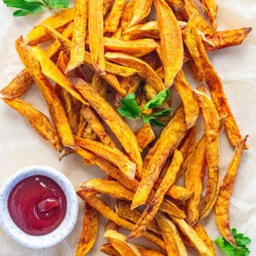 A birds-eye view of crispy air fryer sweet potato fries on a piece of parchment paper with parsley scattered on top and a side of ketchup.