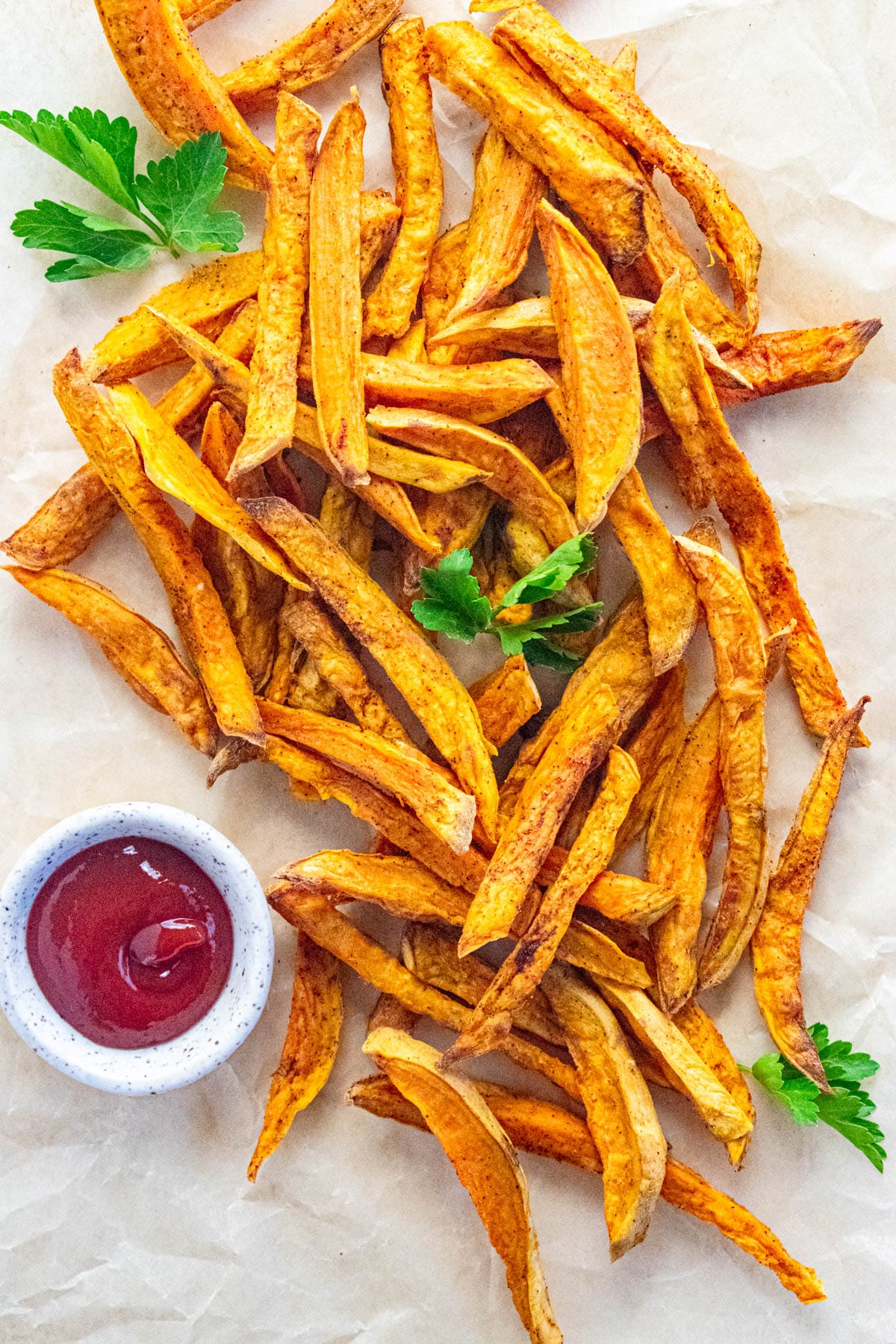 A birds-eye view of crispy air fryer sweet potato fries on a piece of parchment paper with parsley scattered on top and a side of ketchup.