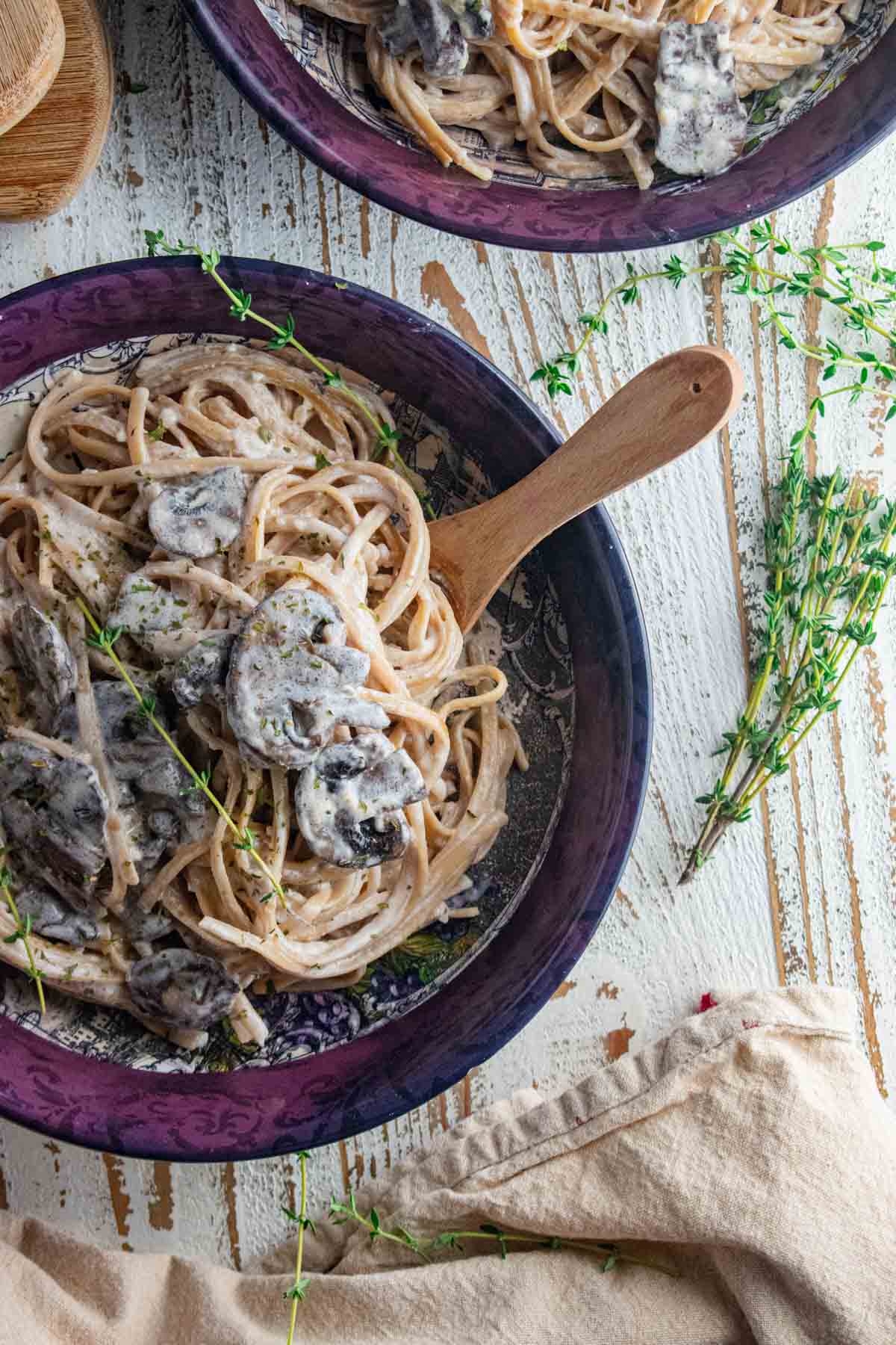 creamy mushroom pasta in a maroon bowl on a white wooden board with a cream colored napkin thrown in