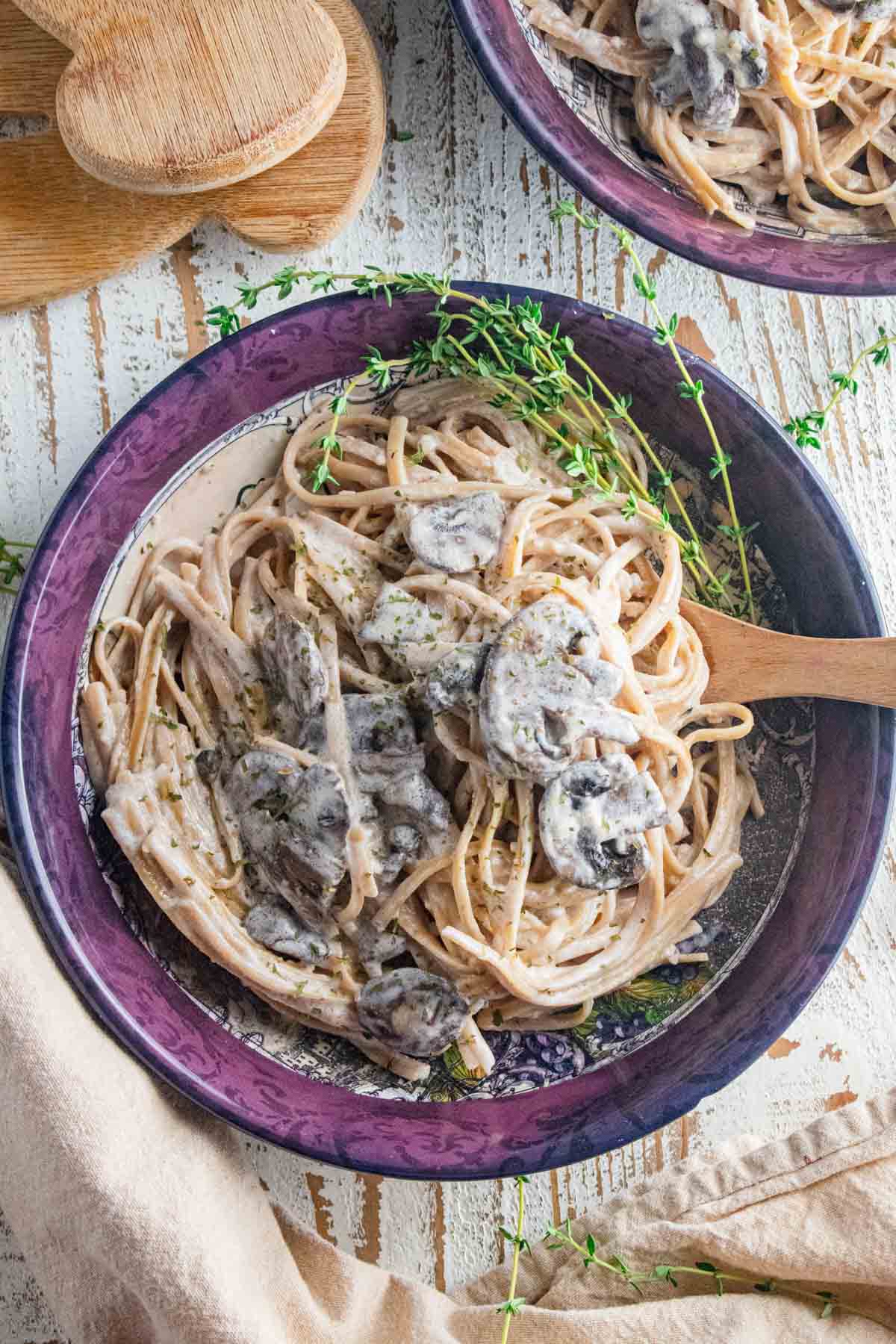 Mushroom pasta in a maroon dish with a wooden spoon slotted in on a white wooden table.