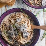 A birds eye view of creamy mushroom pasta in a maroon dish with a wooden slotted spoon on the side and some fresh thyme scattered around.