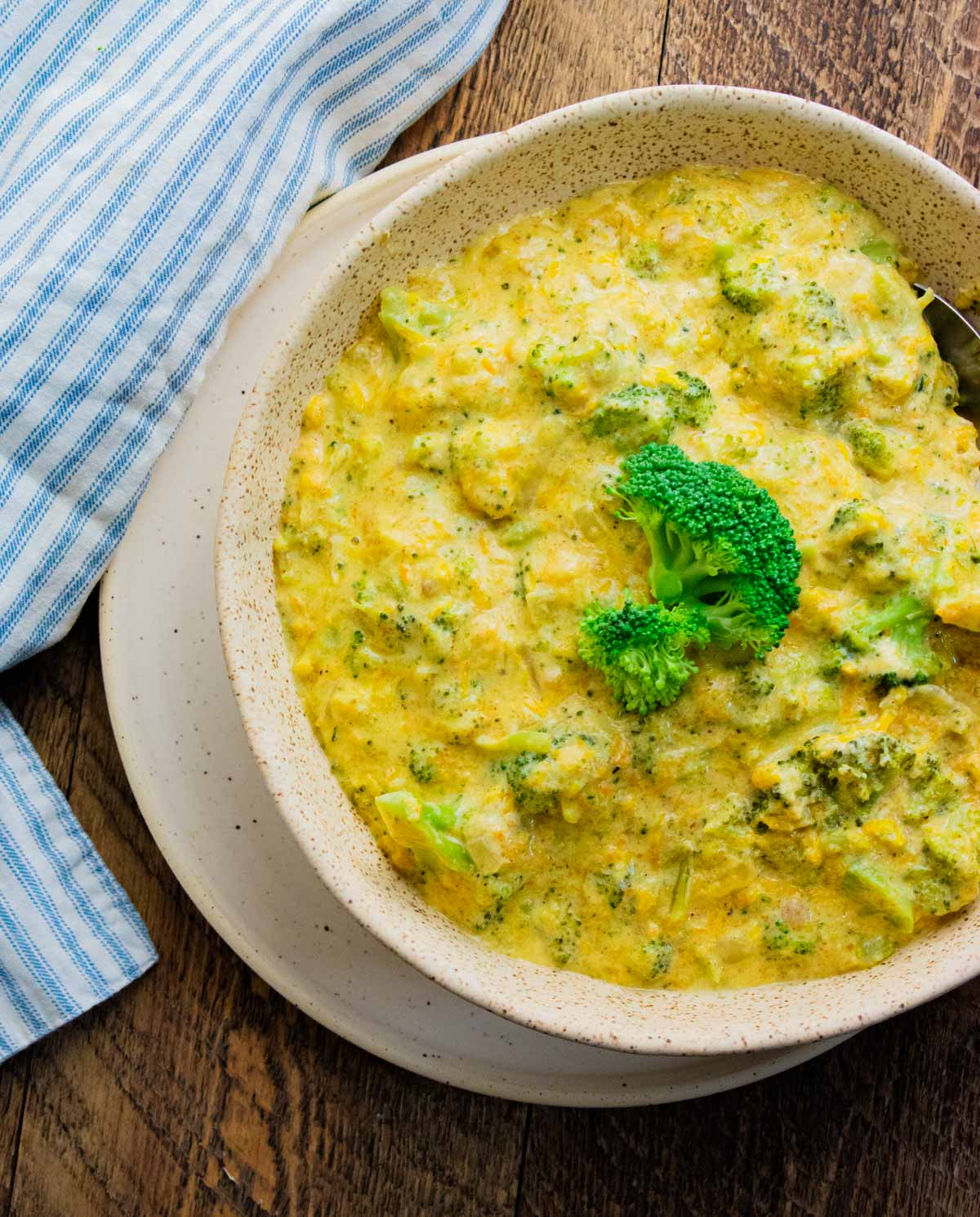 homemade broccoli cheddar soup in a white bowl sitting on a wooden table with a white and blue stripped napkin