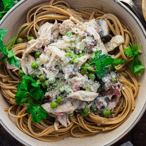 Creamy chicken carbonara in a cream colored bowl with parsley scattered about.