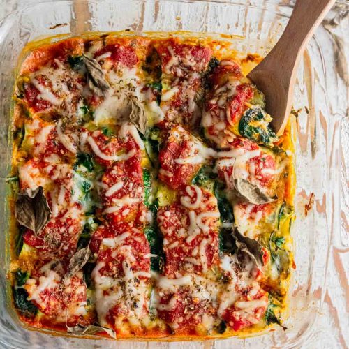 birds eye view of zucchini roll ups in a glass dish with a wooden slotted spoon stuck in the side of the meal