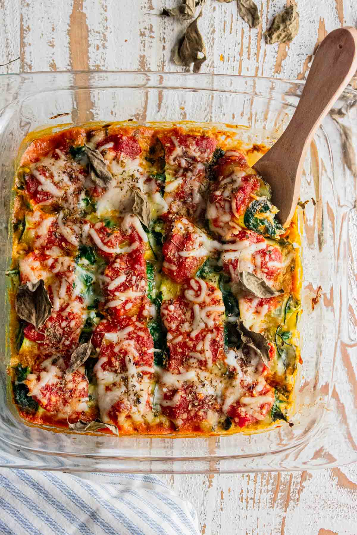 birds eye view of zucchini roll ups in a glass dish with a wooden slotted spoon stuck in the side of the meal