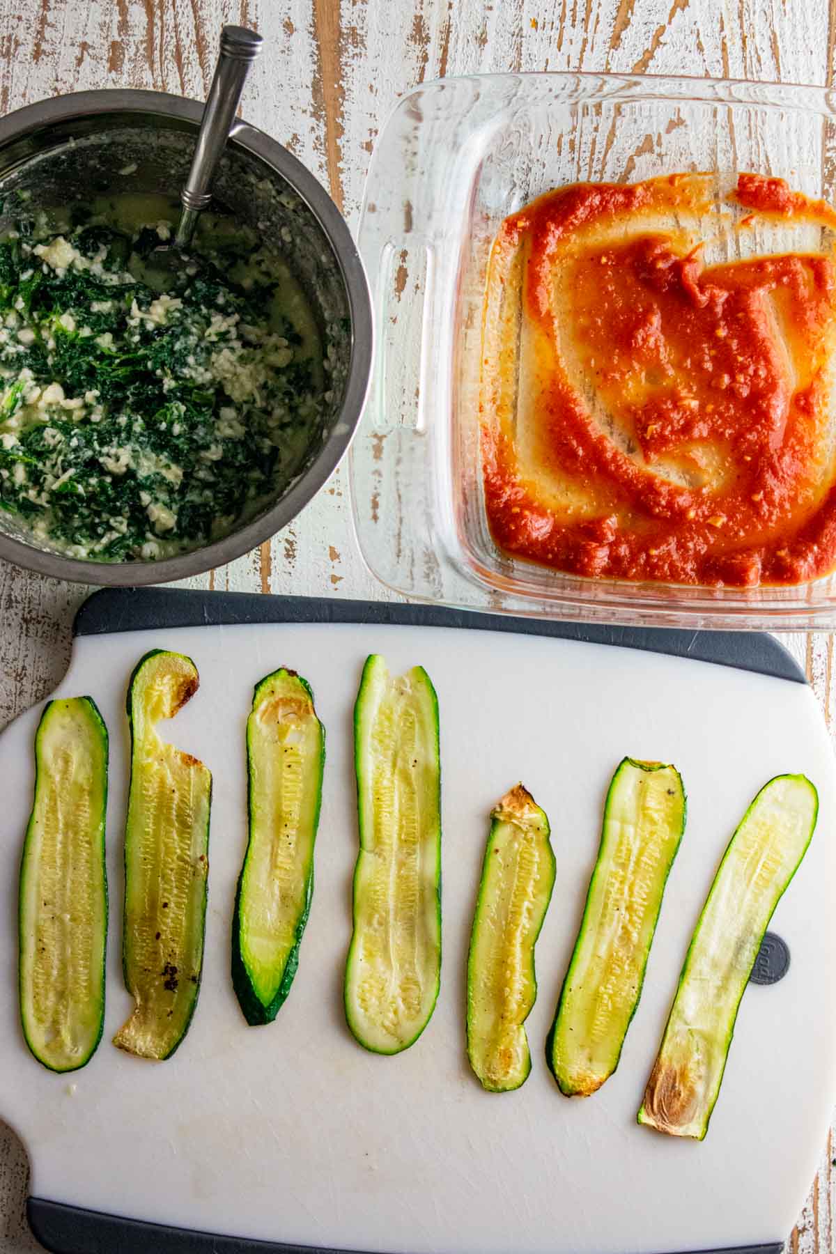 thinly sliced zucchini strips on a cutting board with a bowl of the cottage cheese and spinach filling and a dish coated with marinara sauce on the bottom