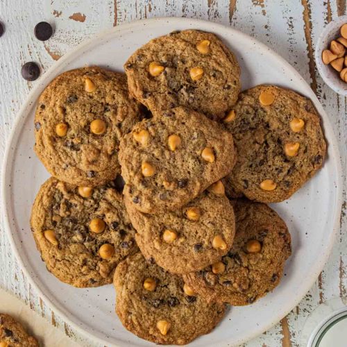 A plate of butterscotch chocolate chip cookies on a white wooden table with butterscotch and chocolate chip morsels scattered around.