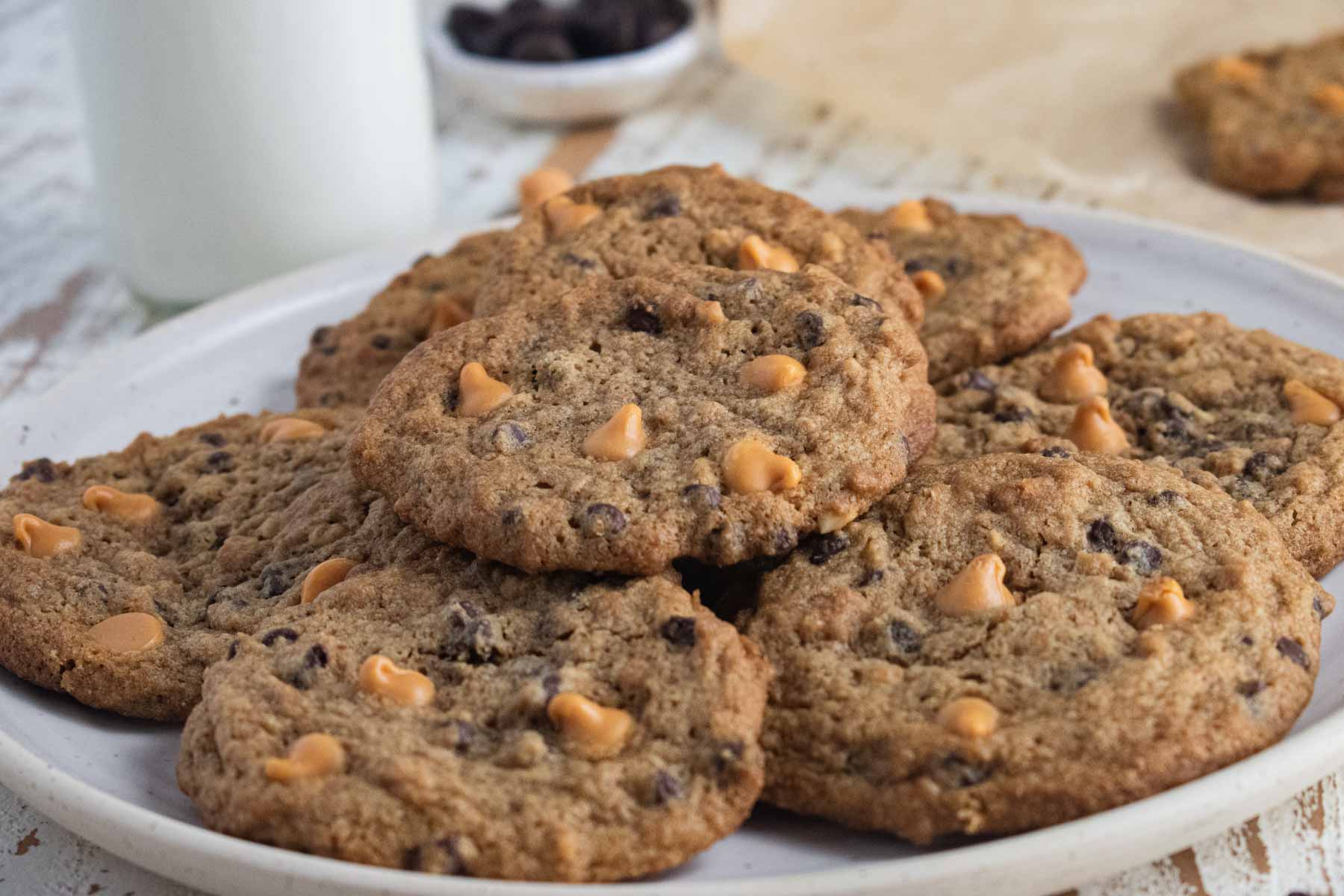 A close up of a plate of butterscotch chocolate chip cookies on a white plate with a glass of milk in the background.