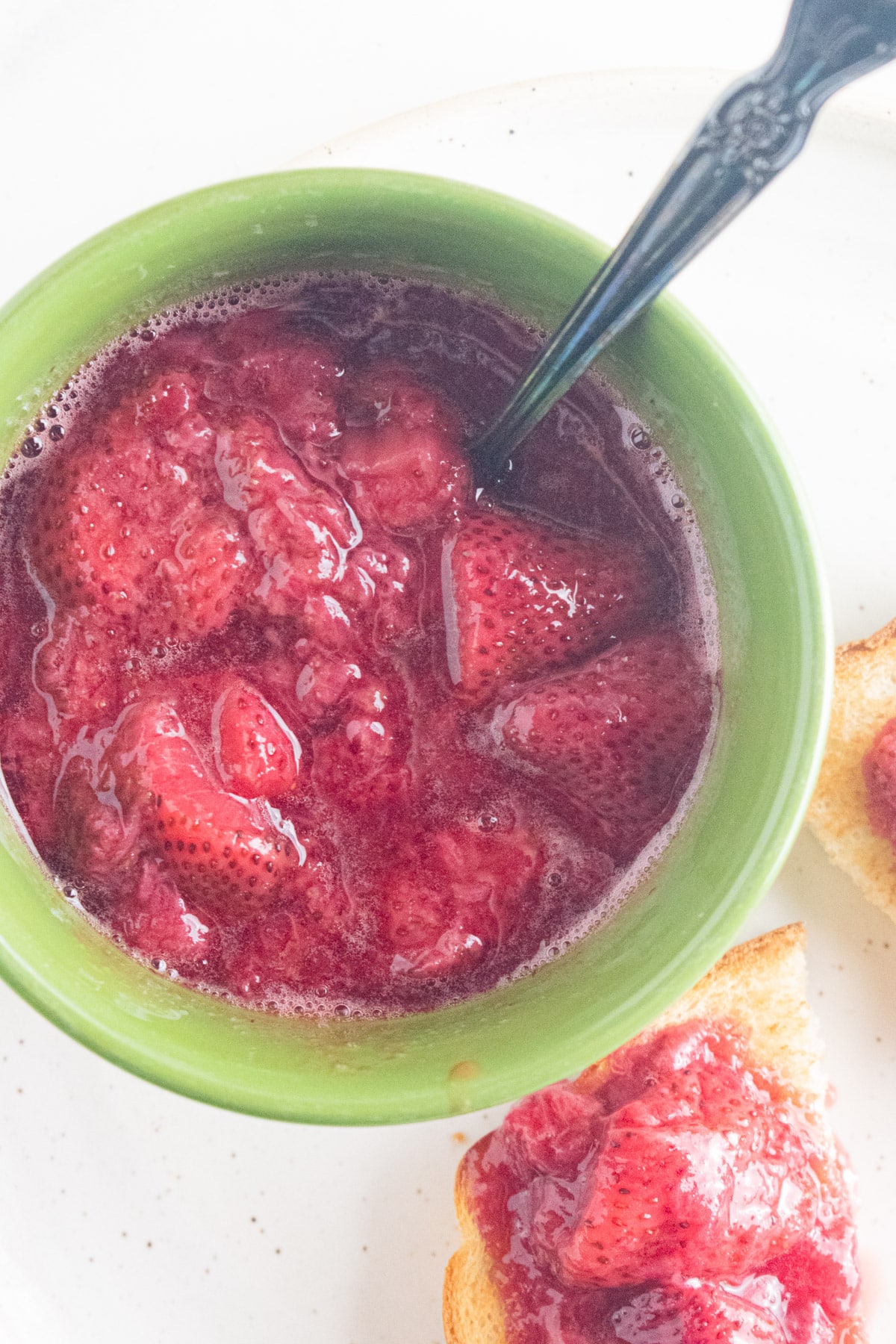 A close up of strawberry compote in a green bowl with a slotted spoon.