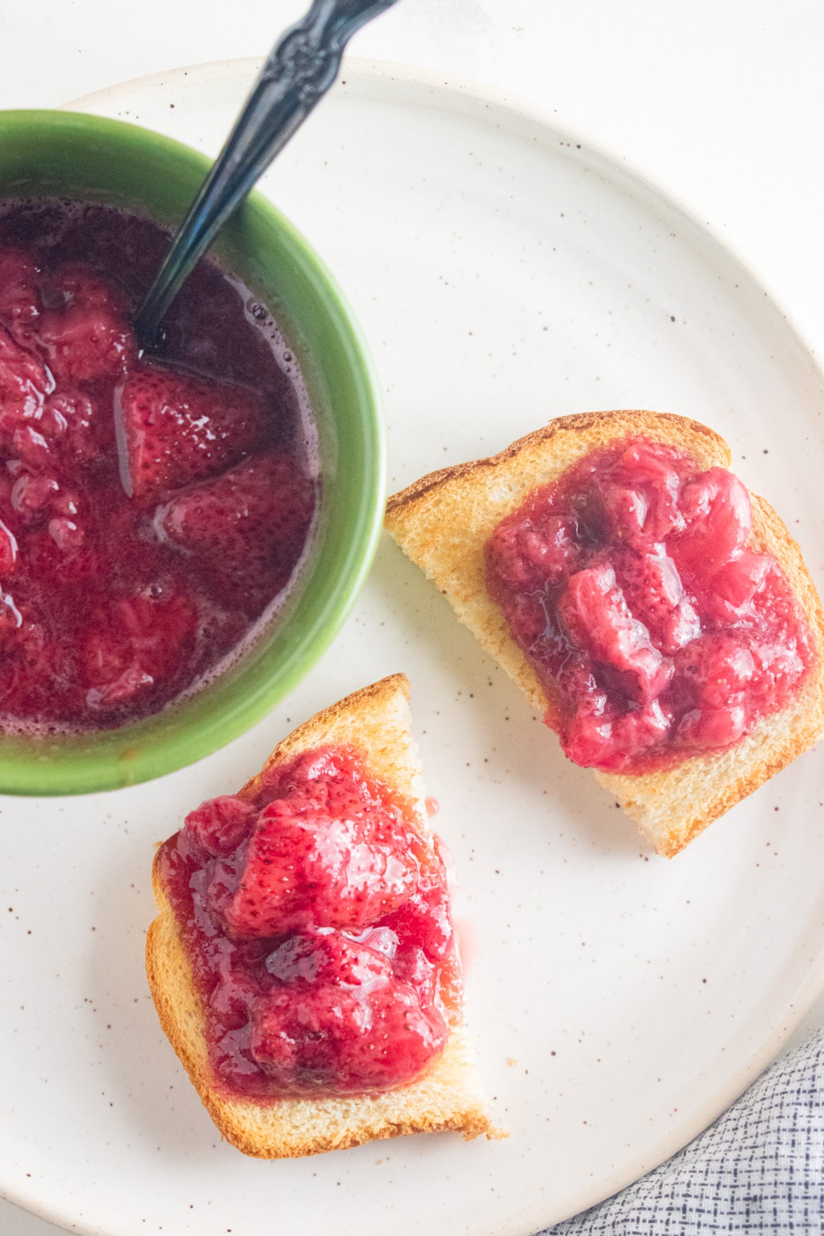 Strawberry compote in a green bowl with a slotted spoon and spread on toast.