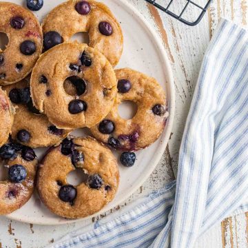 Blueberry donuts on a cream colored plate with a cooling rack in the top right corner and a blue and white checkered napkin laid on the bottom.