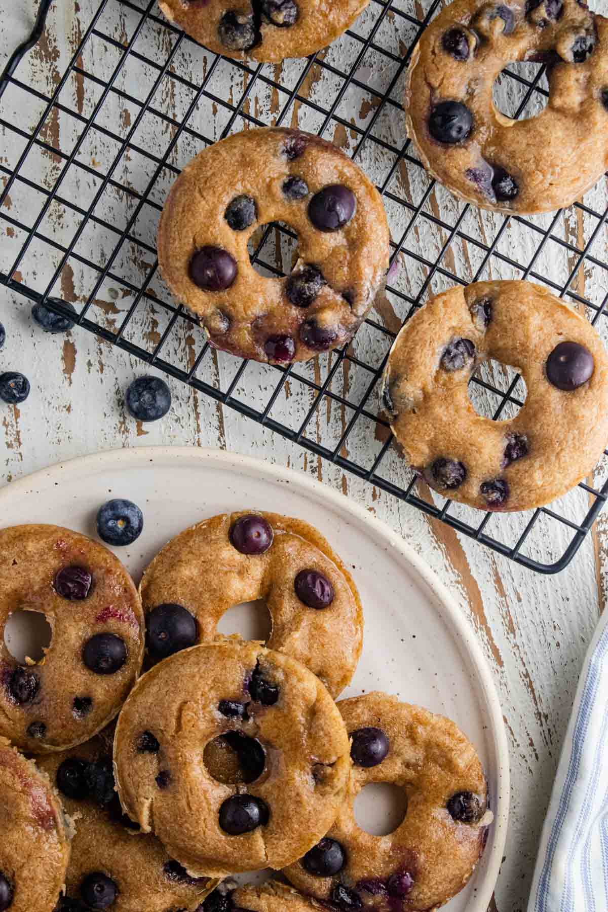 Healthy blueberry donuts on top of a cream colored plate and a cooling rack with blueberries scattered around.