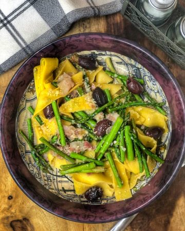 asparagus olives and pancetta paparradelle in a maroon bowl on a wooden board