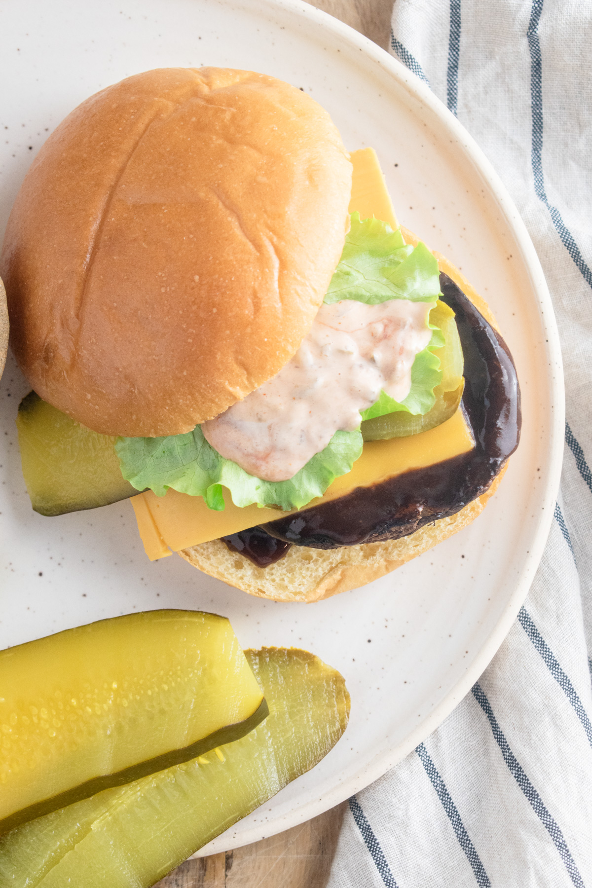 A birds eye view of a portobello mushroom burger on a bun with cheese and a burger sauce and a side of pickles. 