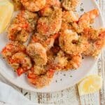 Air fryer shrimp on a white plate on top of a white wooden board with slices of lemon on the side.