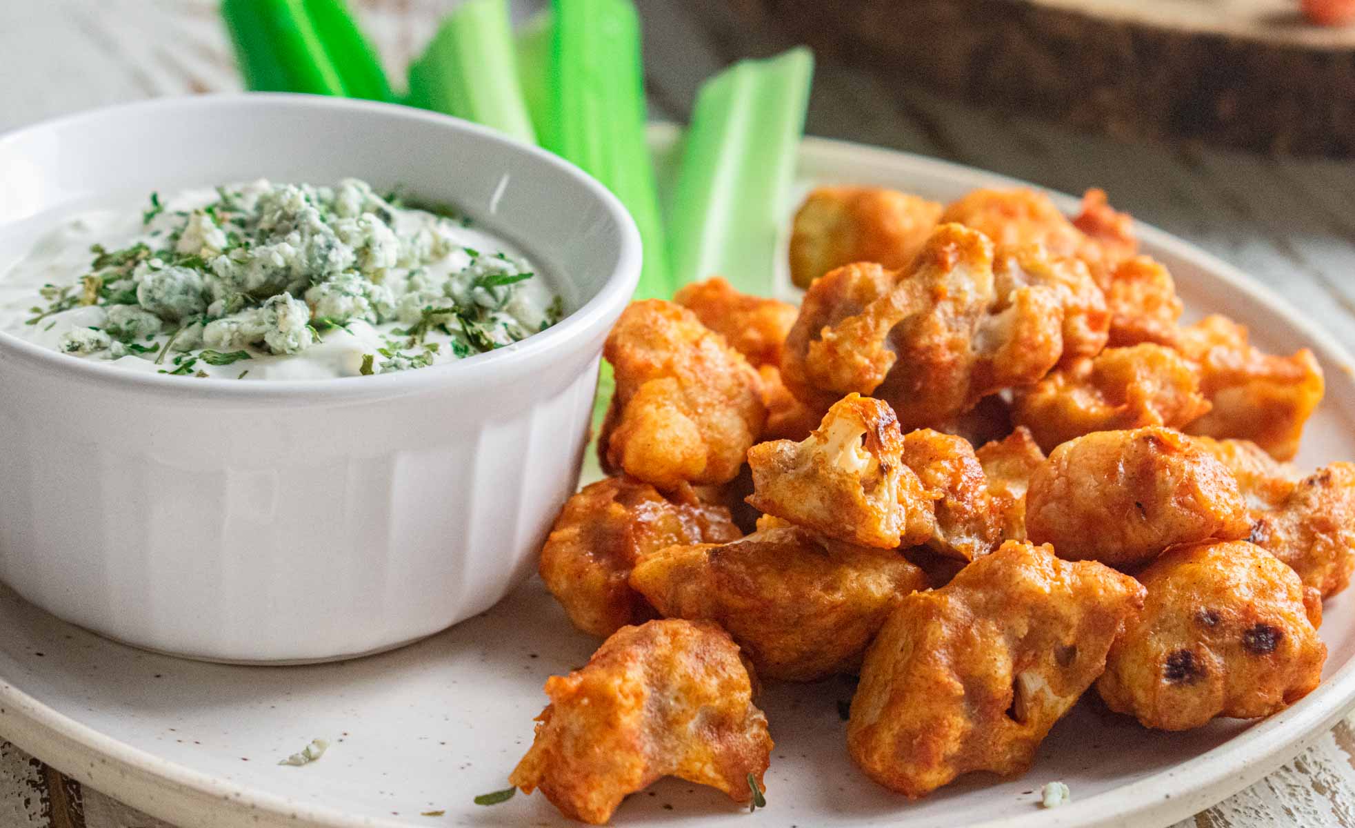 close up of crispy buffalo cauliflower bites on a white plate with blue cheese in a white container and sliced celery stalks