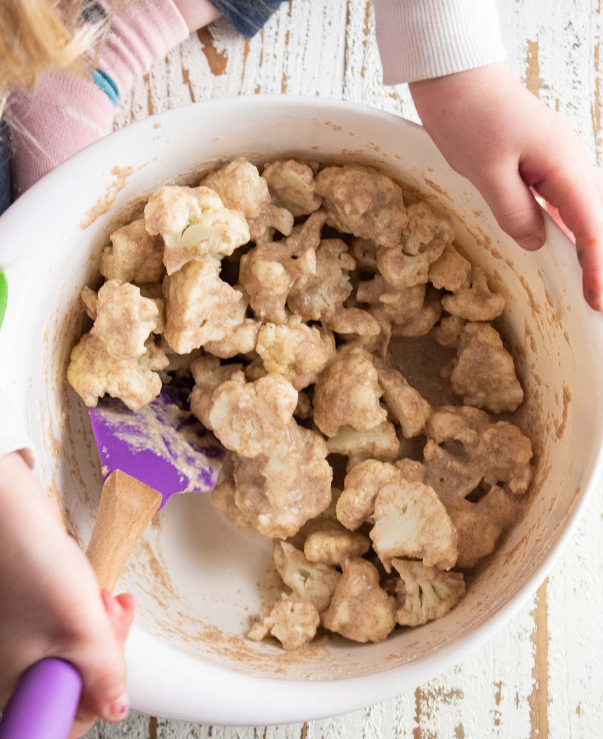 a little child sitting on a table mixing cauliflower with flour using a spatula