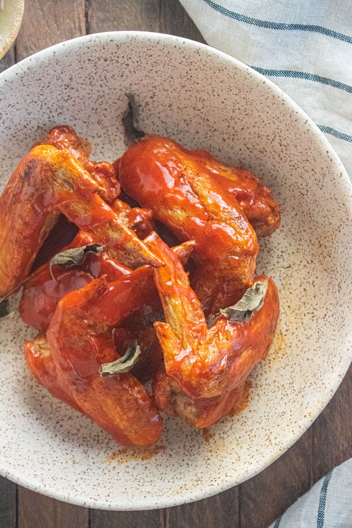 A close up of the crispy buffalo wings on a cream colored plate on top of a wooden table with a striped napking next to the bowl.