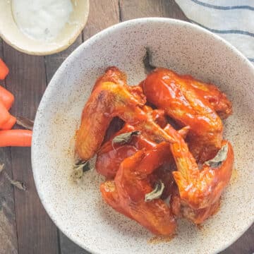 Buffalo wings in a white bowl on a wooden table with carrots laid down next to them and a stripped napkin.