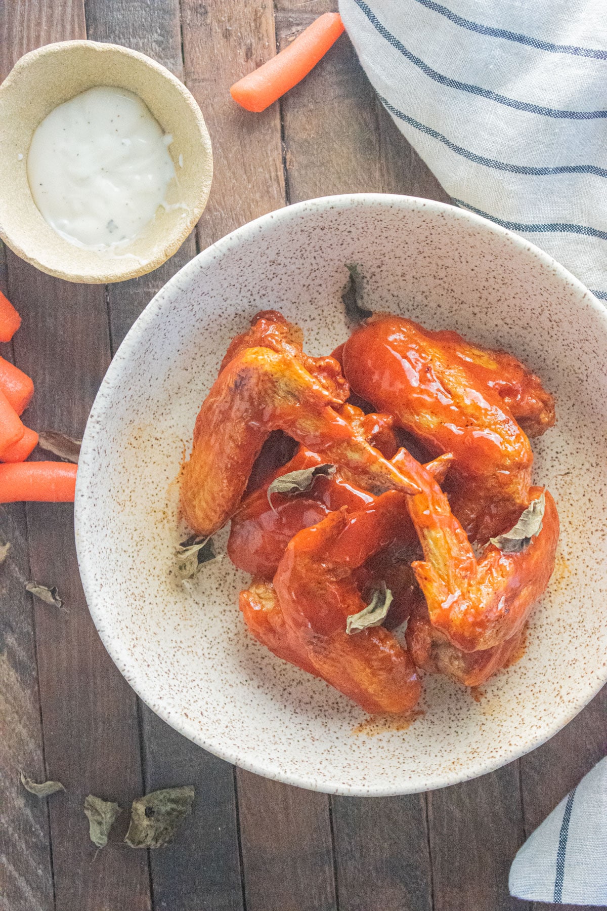 Buffalo wings in a white bowl on a wooden table with carrots laid down next to them and a stripped napkin.