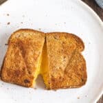 Two crispy pieces of bread on a white plate with oozing cheese in between the two pieces of bread.