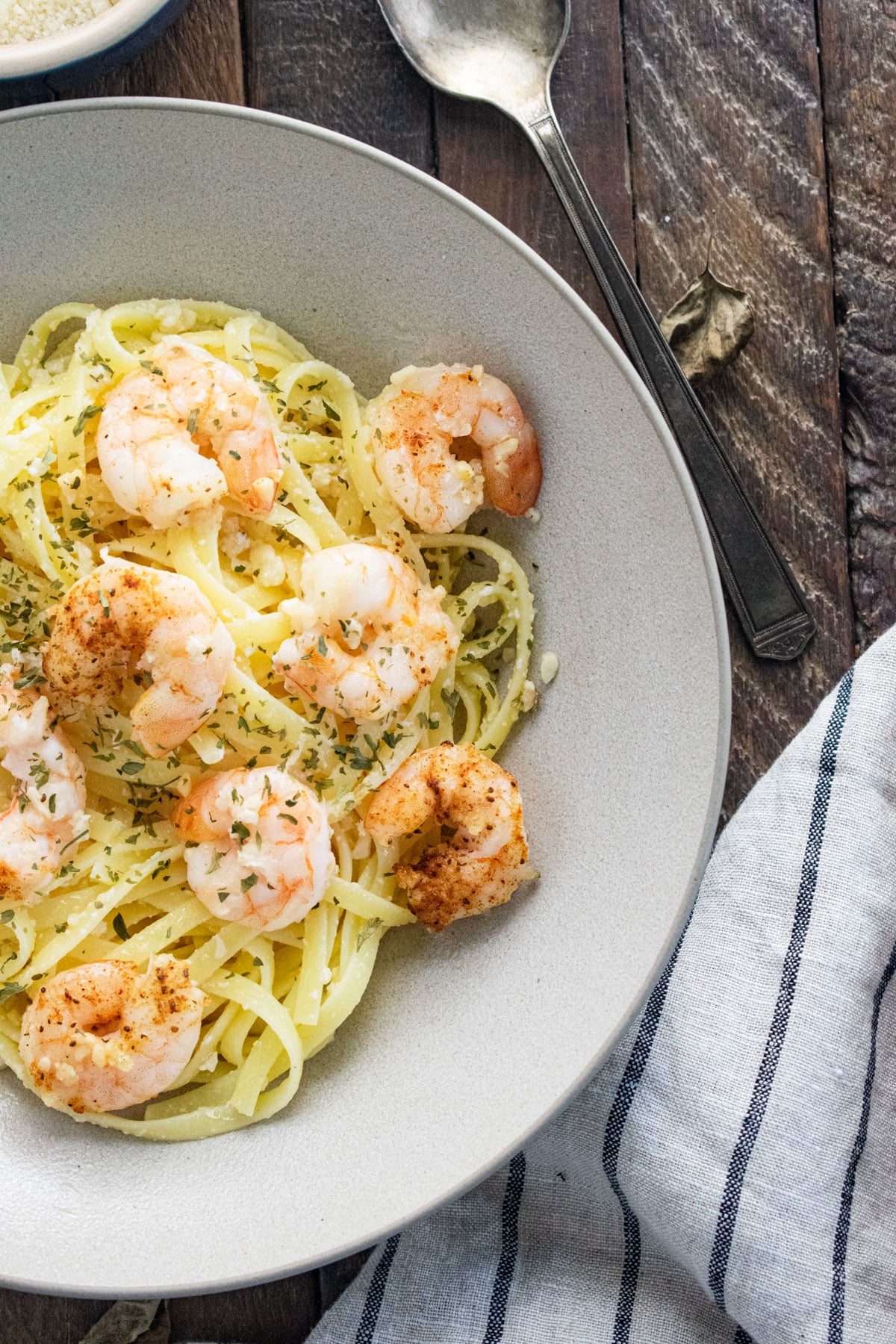 garlic butter shrimp pasta in a white bowl on a wooden table with a striped colored napkin and a silver spoon.