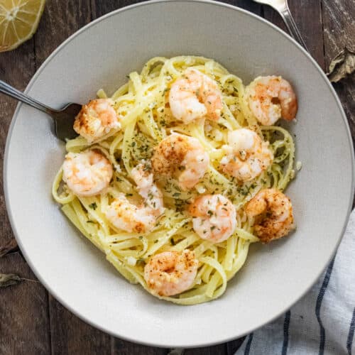 Fettucine and shrimp in a white bowl topped with grated parmesan and a slotted spoon on the side.