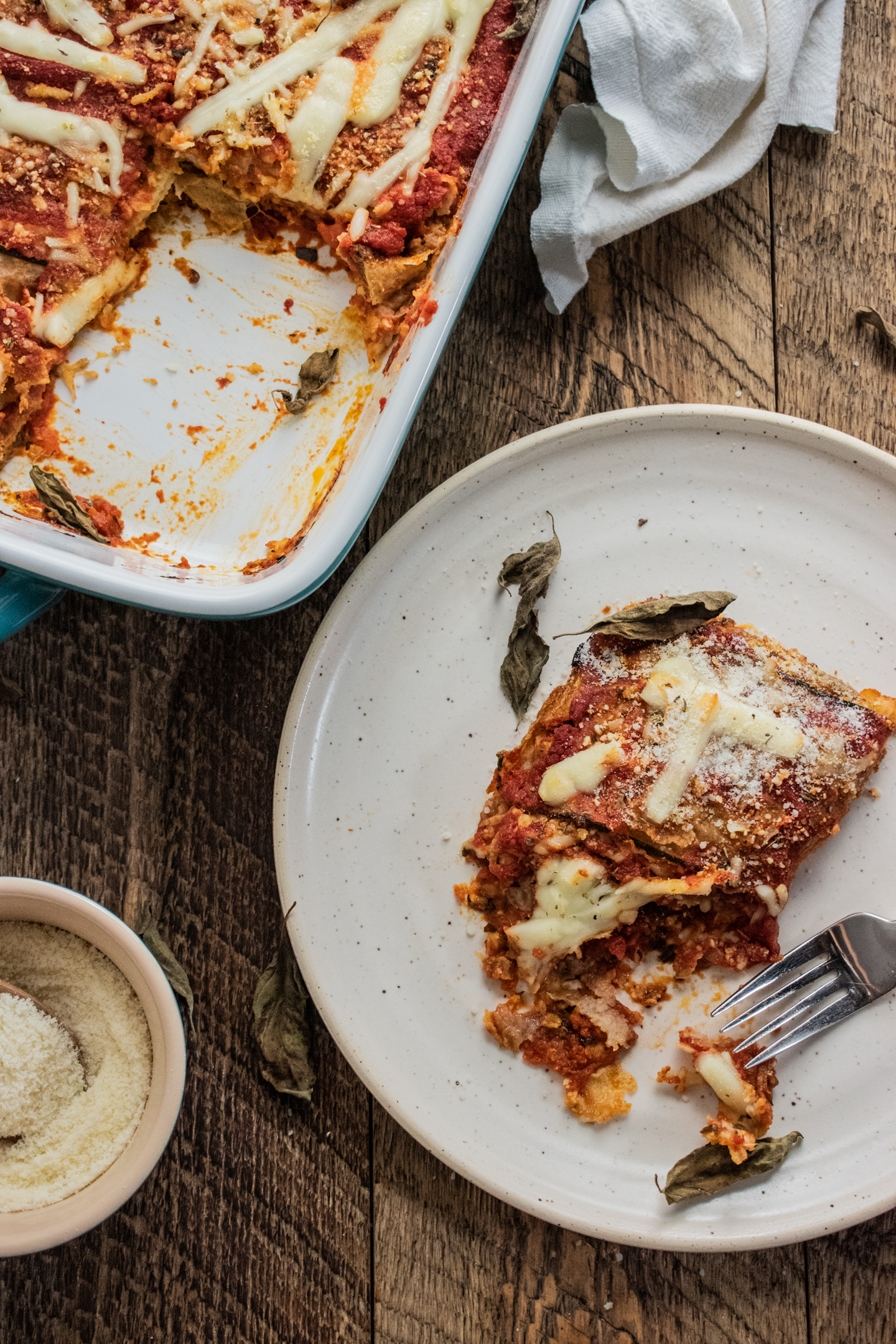 zucchini parmesan in a 9 x 13 inch baking dish on a wooden table with a piece cut out on a white plate next with a fork