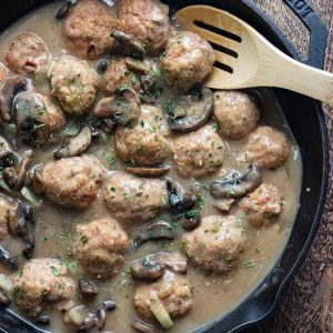 swedish meatlballs stroganoff in a creamy mushroom sauce cooking in a cast iron skillet being stirred by a wooden spoon