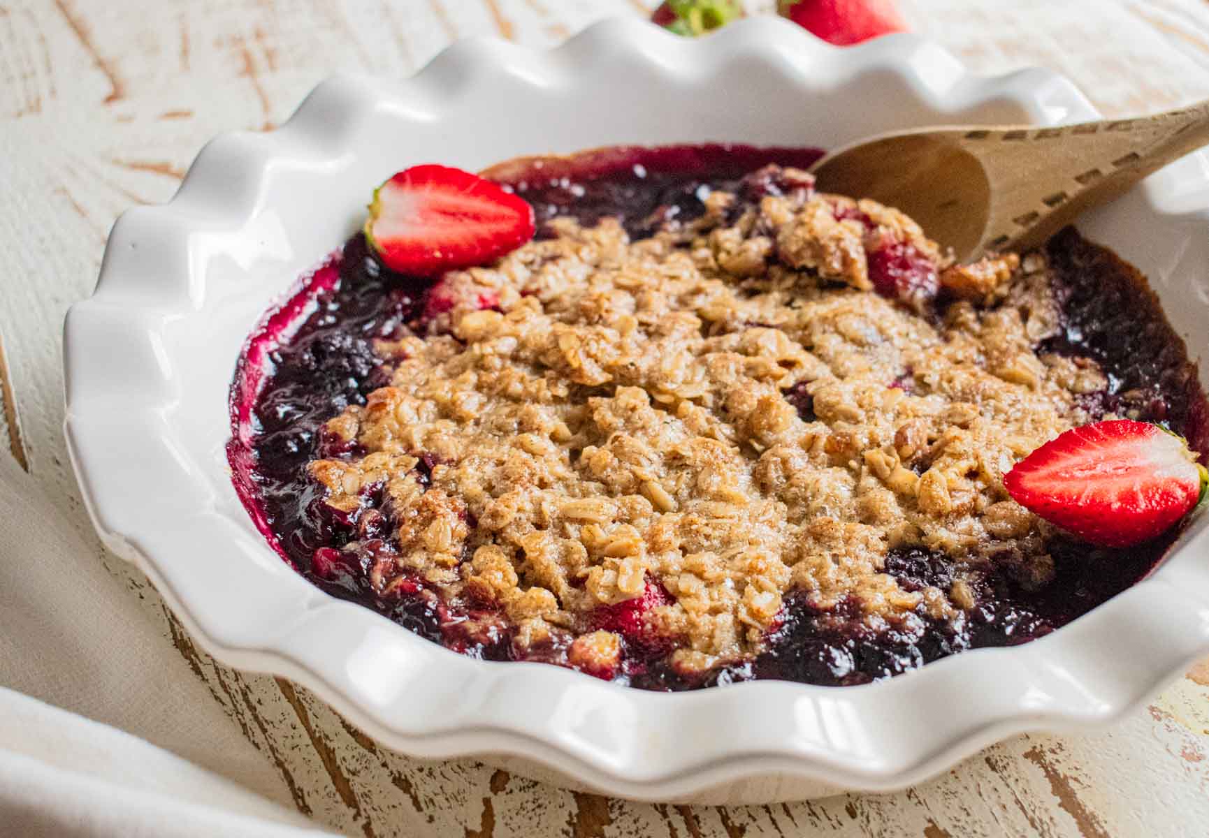 A delicious fruit crisp fresh out of the oven topped with golden brown crumbles in a white pie dish and topped with fresh strawberries. 