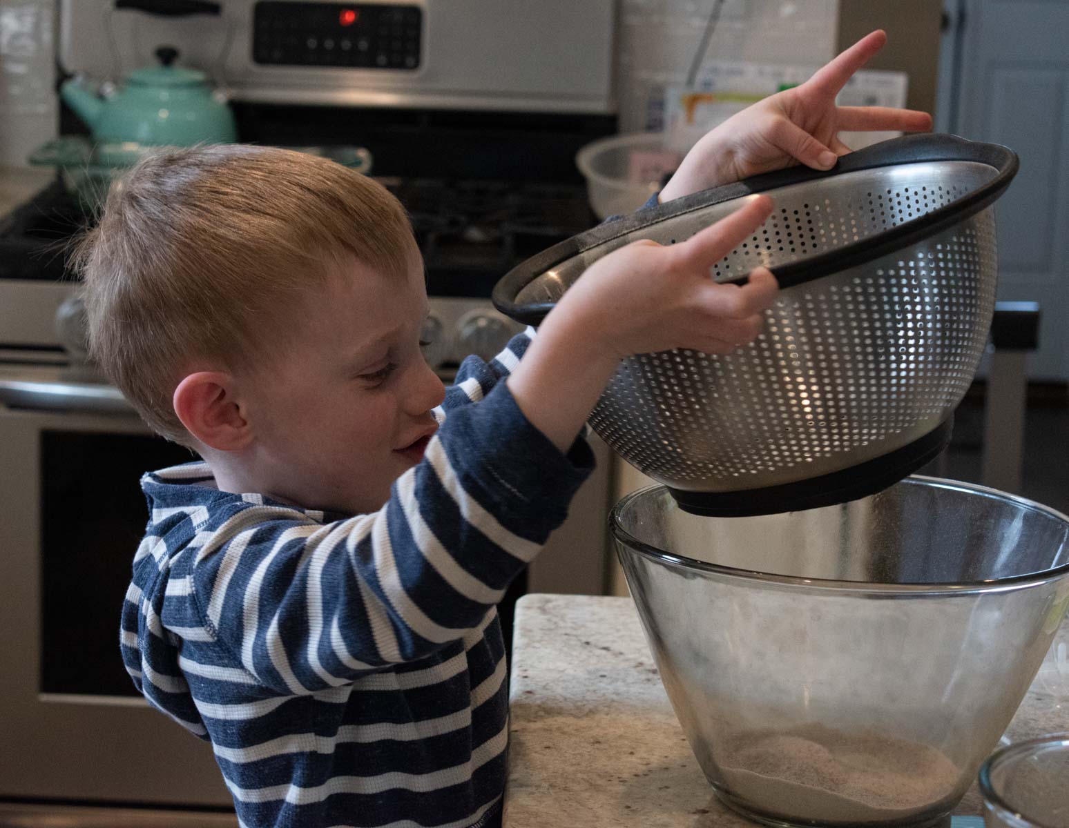 a little blonde boy in the kitchen sifting flour using a colander and a glass bowl. This is step 2 