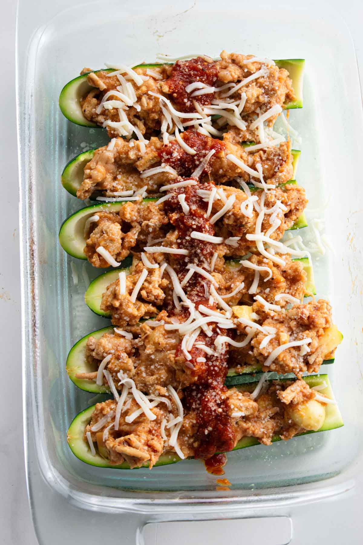 zucchini boats filled with ground chicken mixture and topped with shredded cheese and marinara sauce in a glass baking dish. 