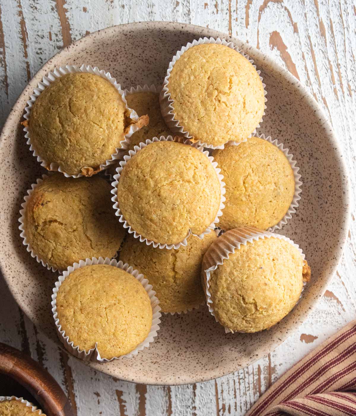 A birds eye view of gluten-free corn muffins in a cream colored dish on a white wooden table next to a striped napkin