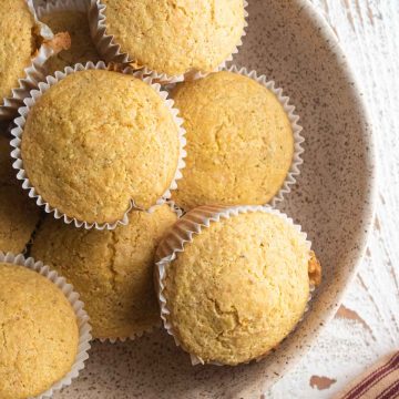 close up gluten-free corn muffins in a cream colored bowl on a white wooden board with a striped napkin next to the bowl