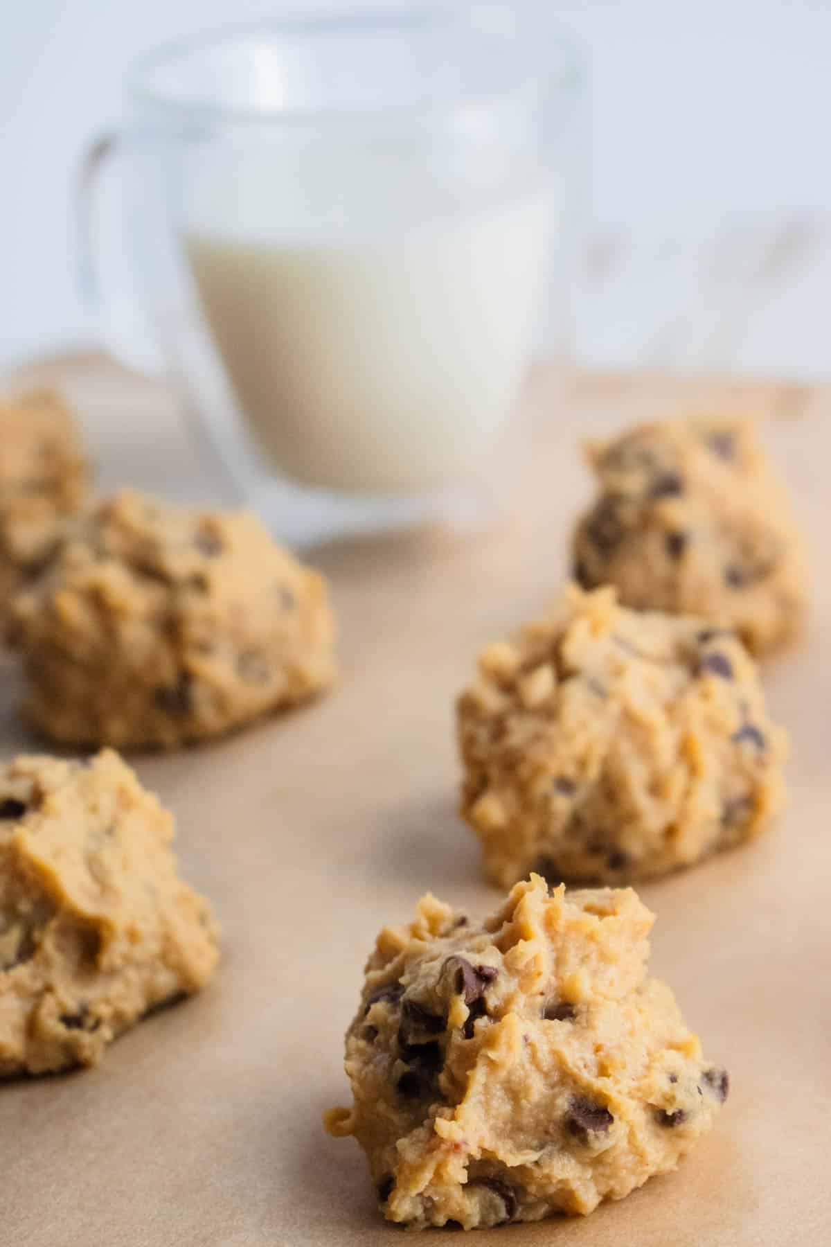 A close up of a cookie dough bite on some parchment paper with more cookie dough bites and a cool glass of milk in the background.